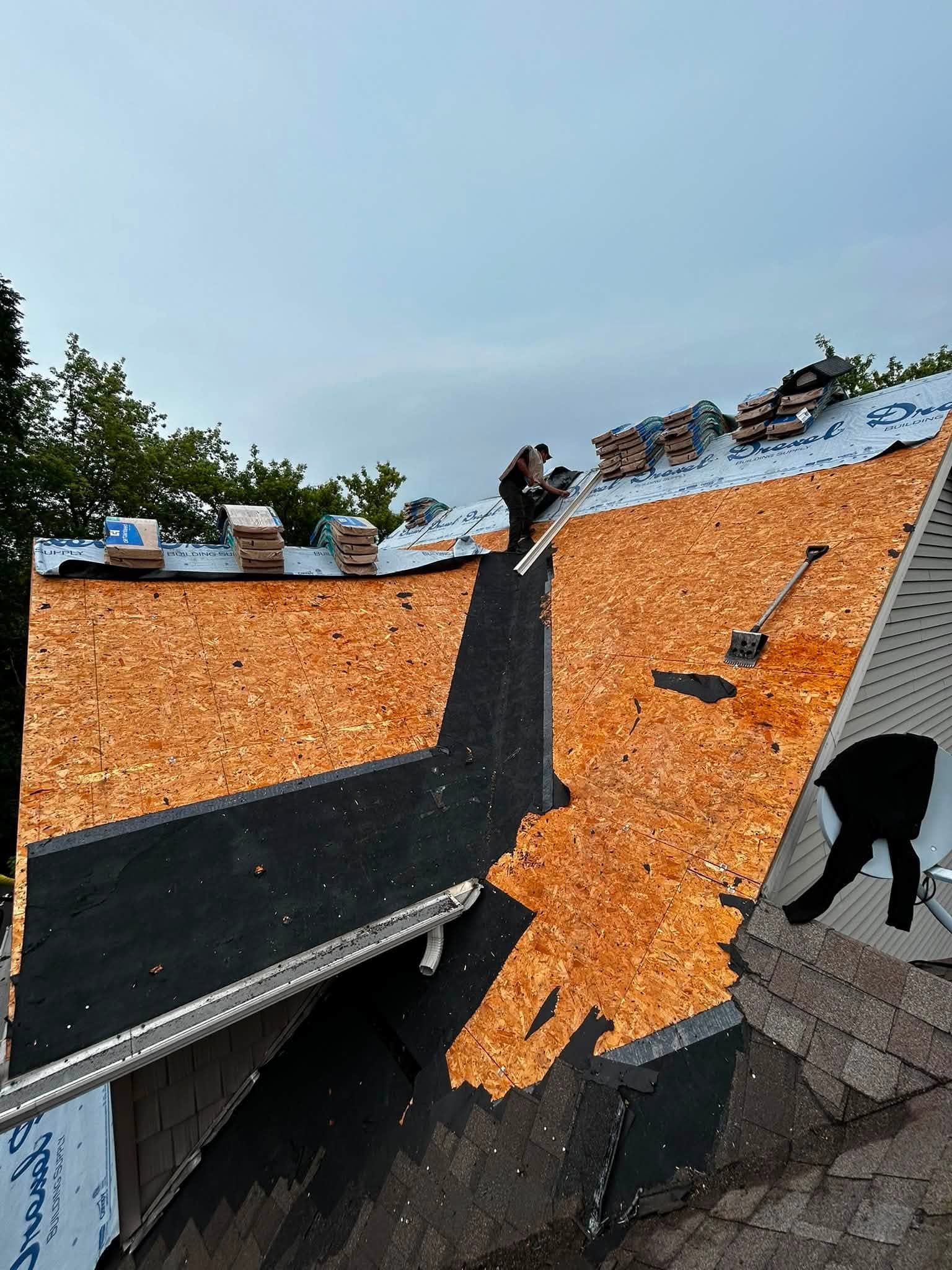 Roofing worker beside exposed decking, underlayment, and staged shingle bundles
